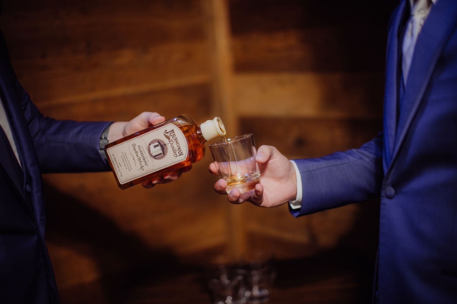 Hands pouring whiskey into glasses during wedding preparation.
