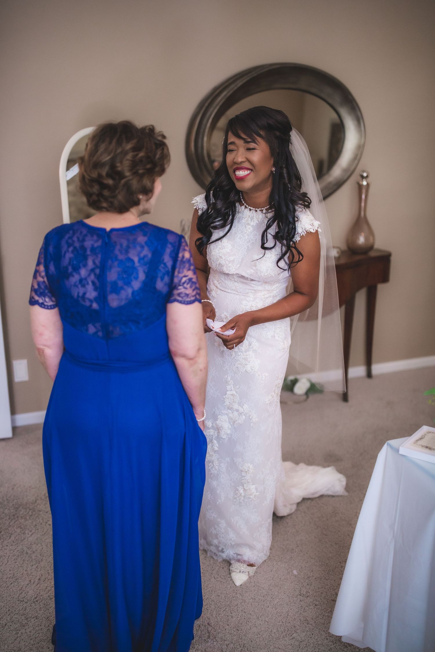 Two women share a happy conversation during wedding preparations in front of an oval mirror.