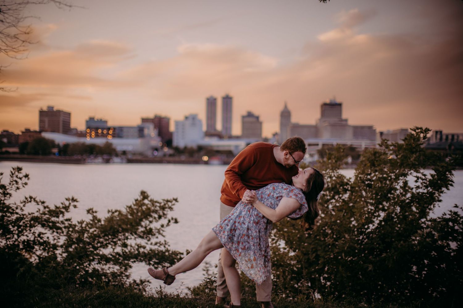 Romantic couple embraces in front of a city skyline at sunset with water and greenery in the foreground.