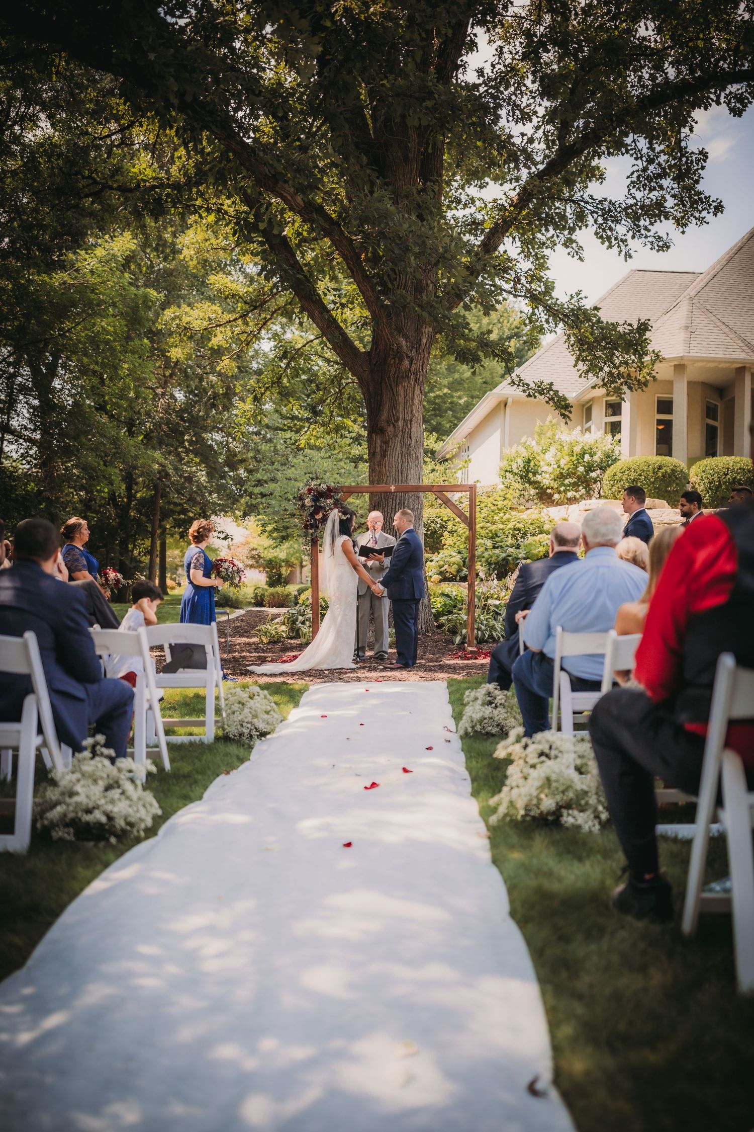 White aisle runner leads through rows of chairs at an outdoor wedding ceremony under tall trees.
