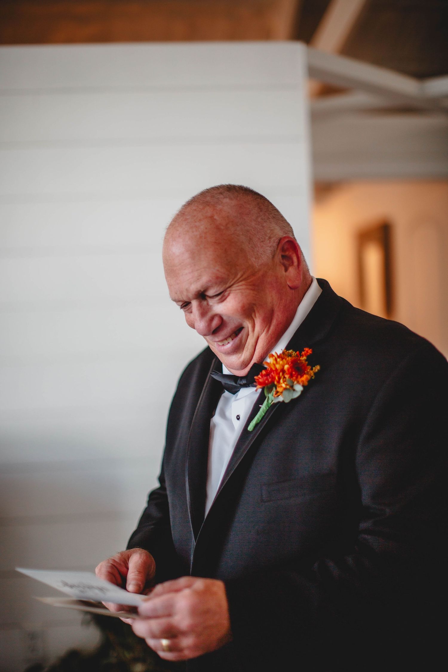 Person in black tuxedo with orange boutonniere reads from papers with joyful expression.