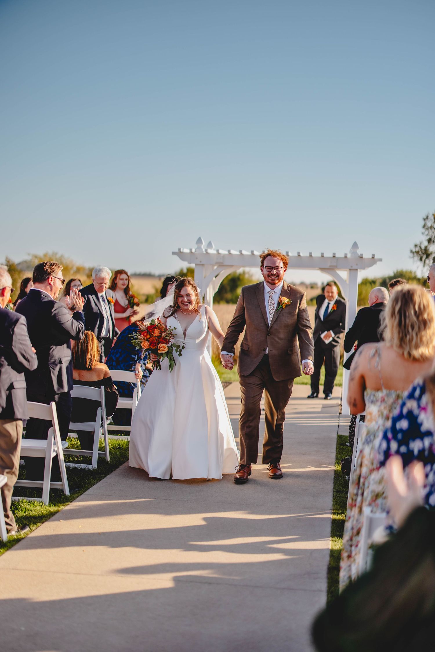 Happy couple walks down aisle after outdoor ceremony with autumn bouquet and brown suit.