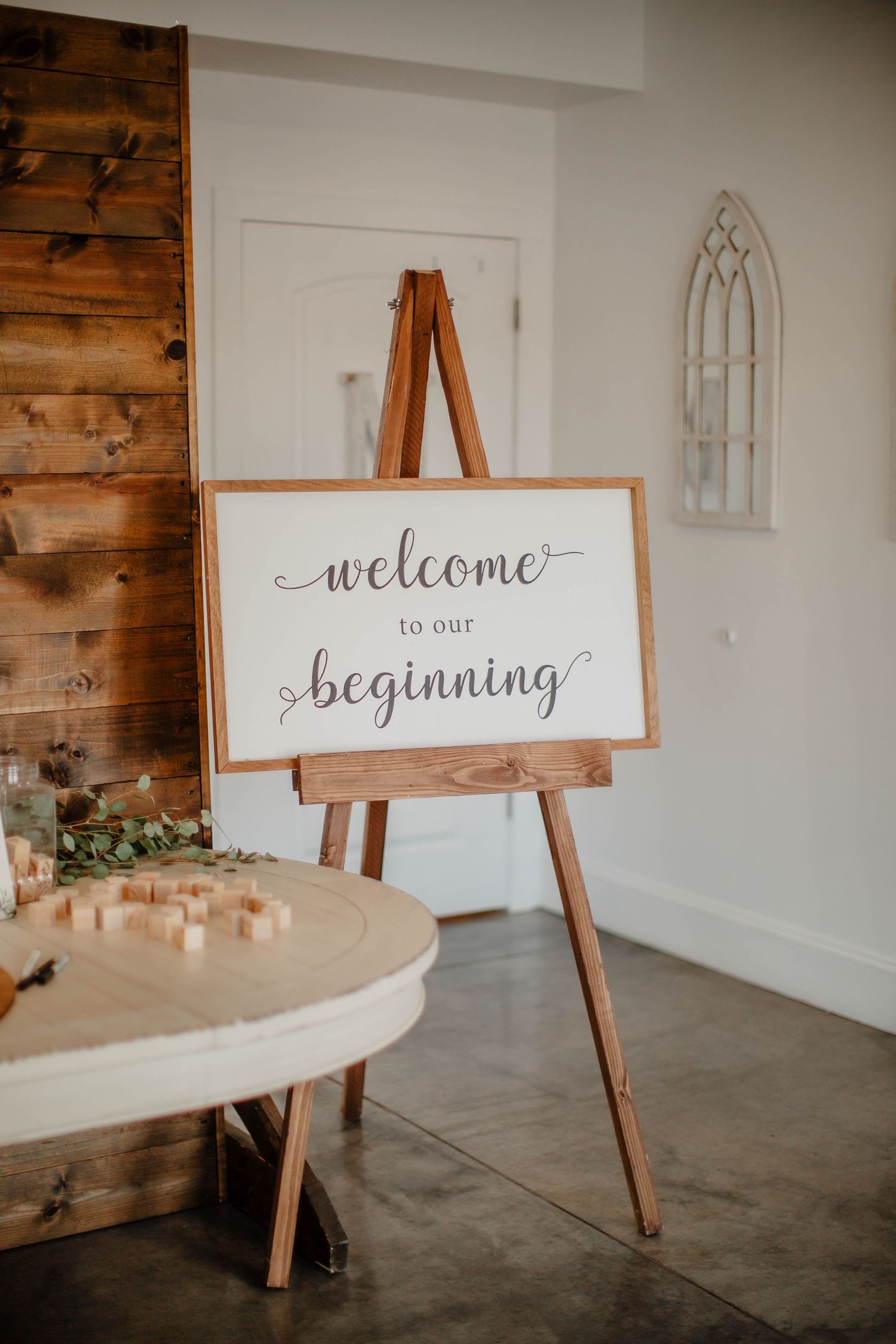 Wooden easel displaying a framed 'welcome to our beginning' sign against a white wall with rustic decor.