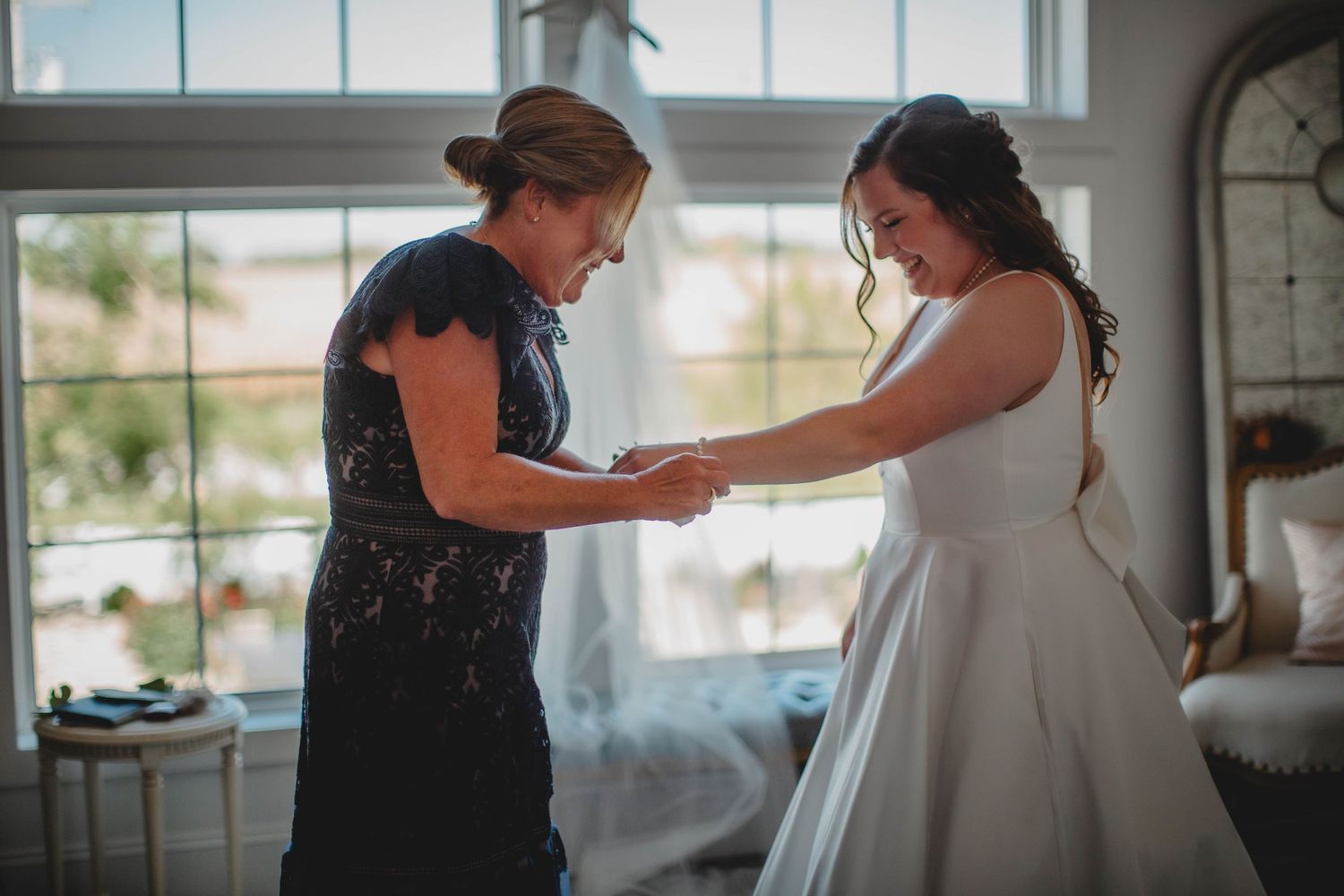 Two people share an emotional moment during wedding dress preparations in a sunlit room.