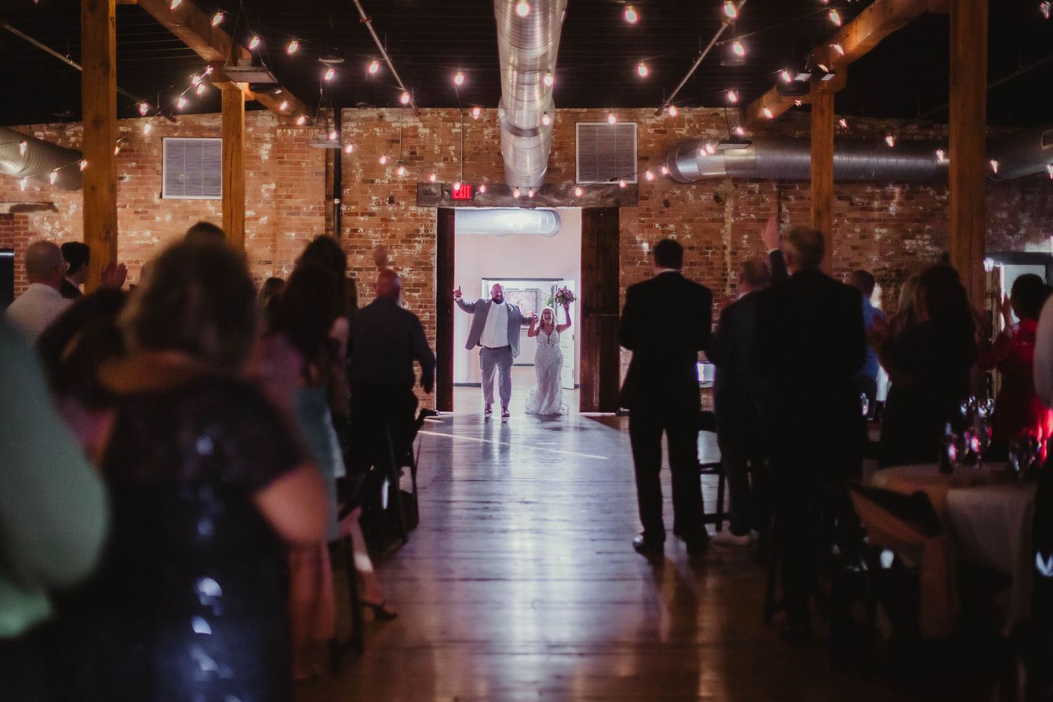 Wedding venue interior showing string lights and exposed brick walls with guests mingling.