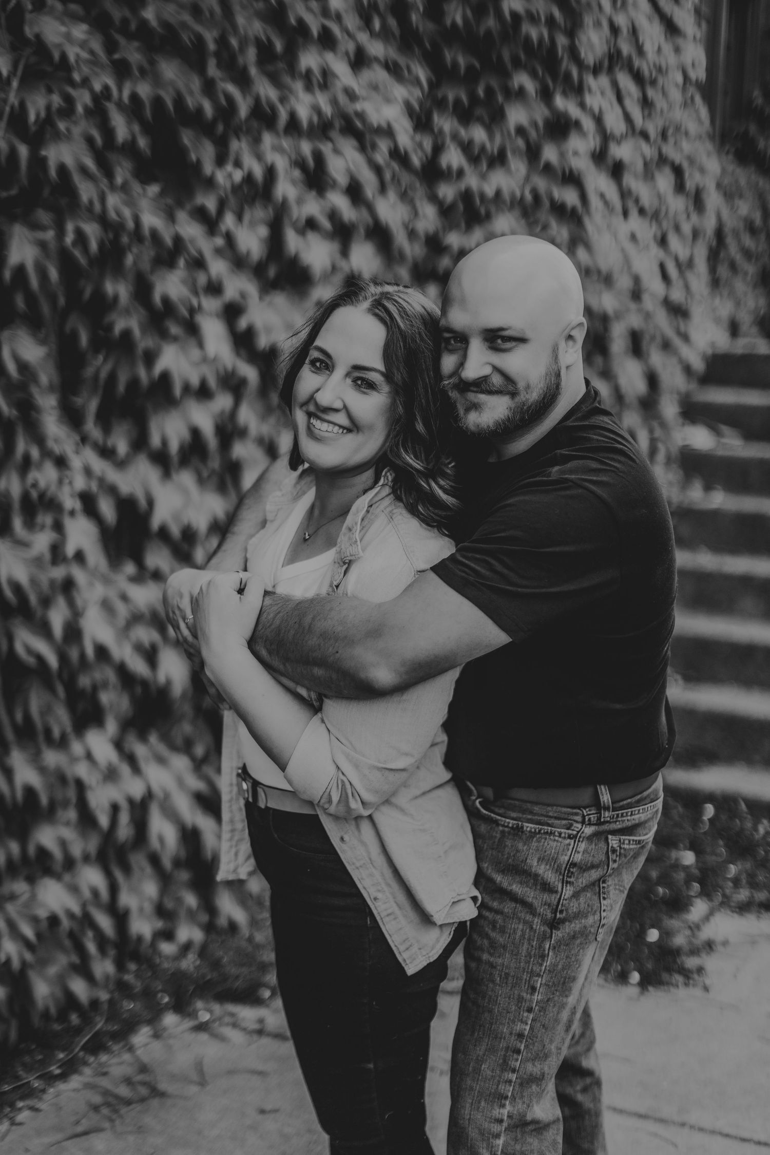 A happy couple embraces while posing in front of a vine-covered wall on a sunny day.