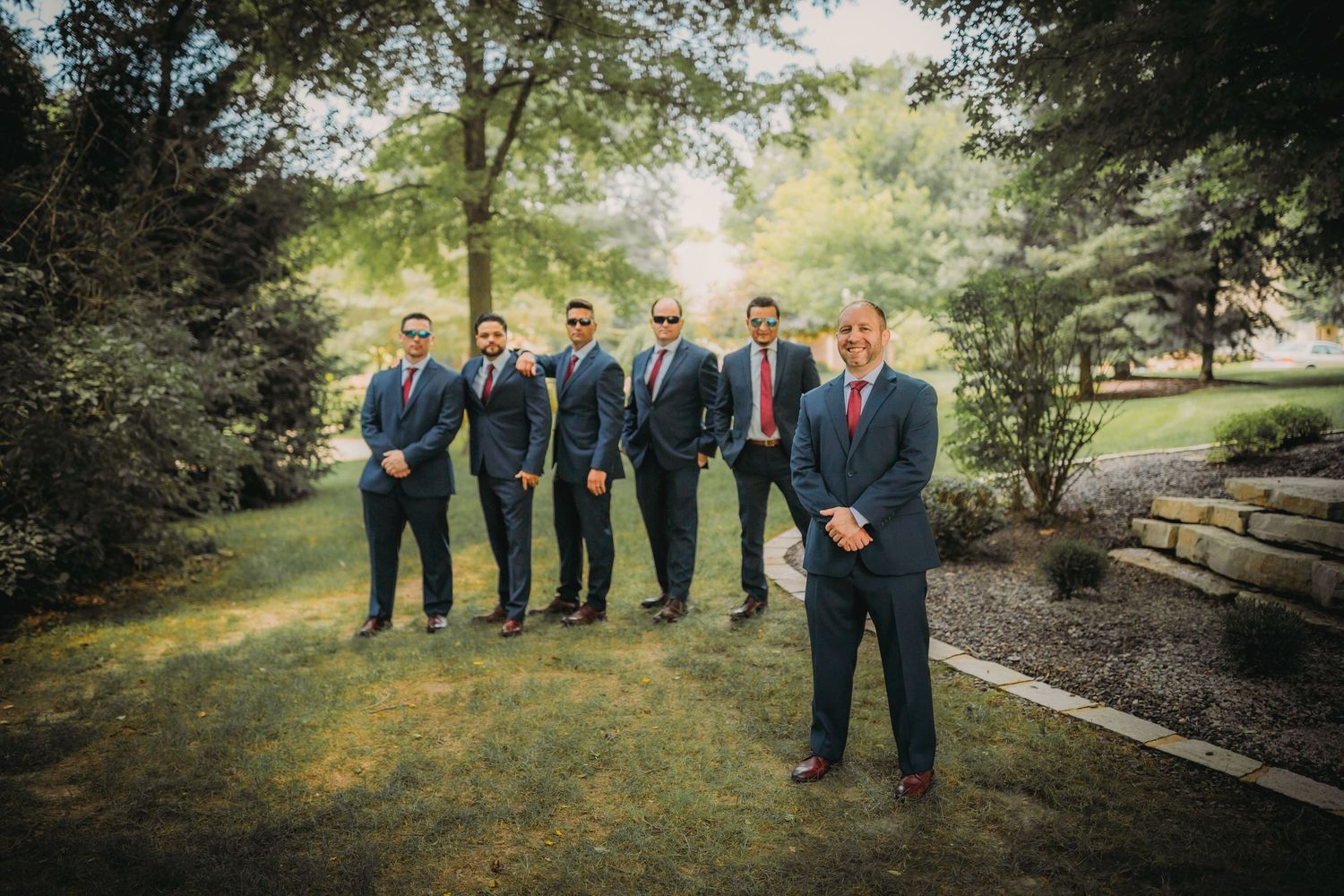 Groomsmen in dark suits pose together on a garden path during sunset.