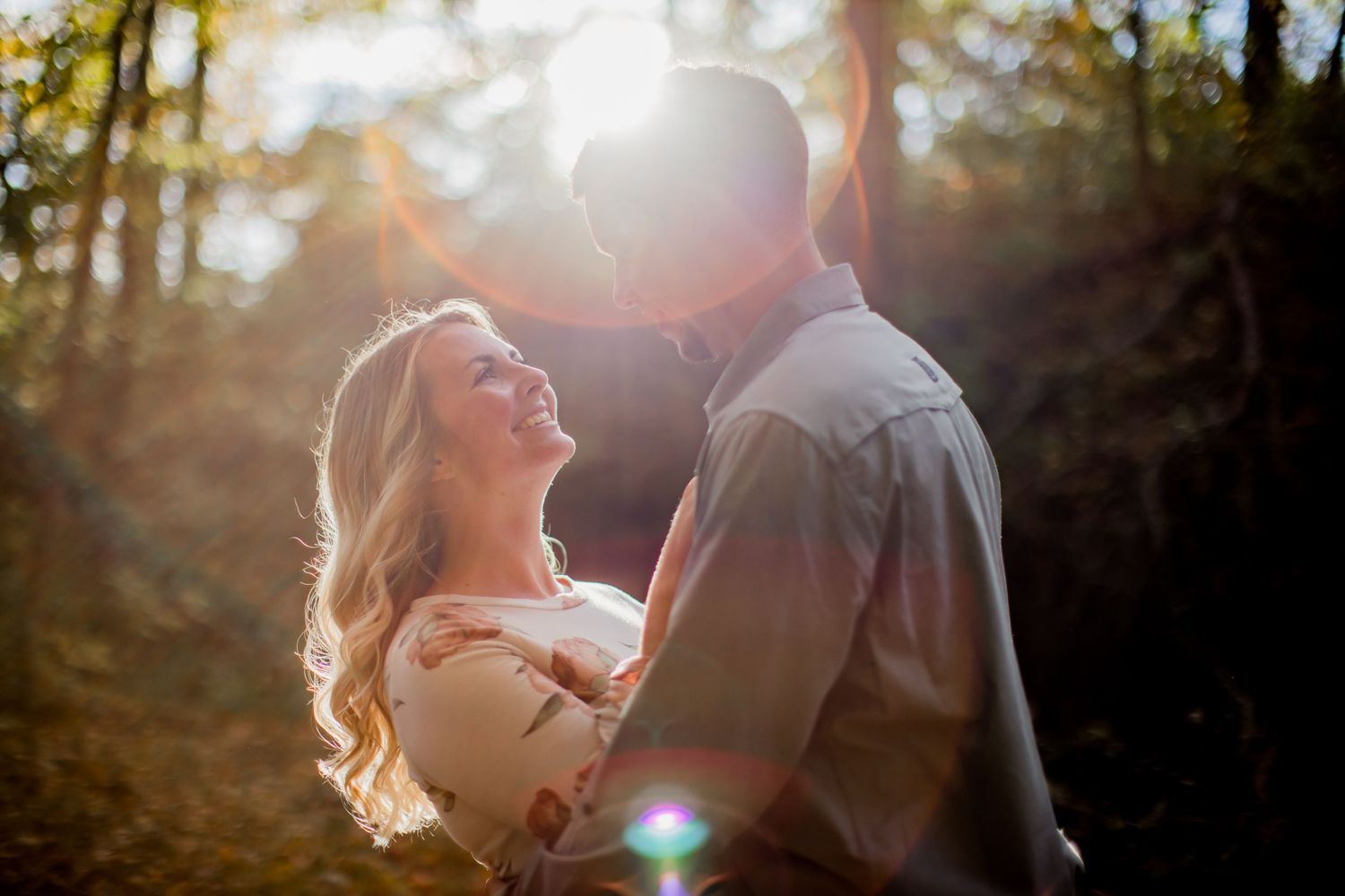 Couple sharing tender moment in backlit forest setting with ethereal sun flares.