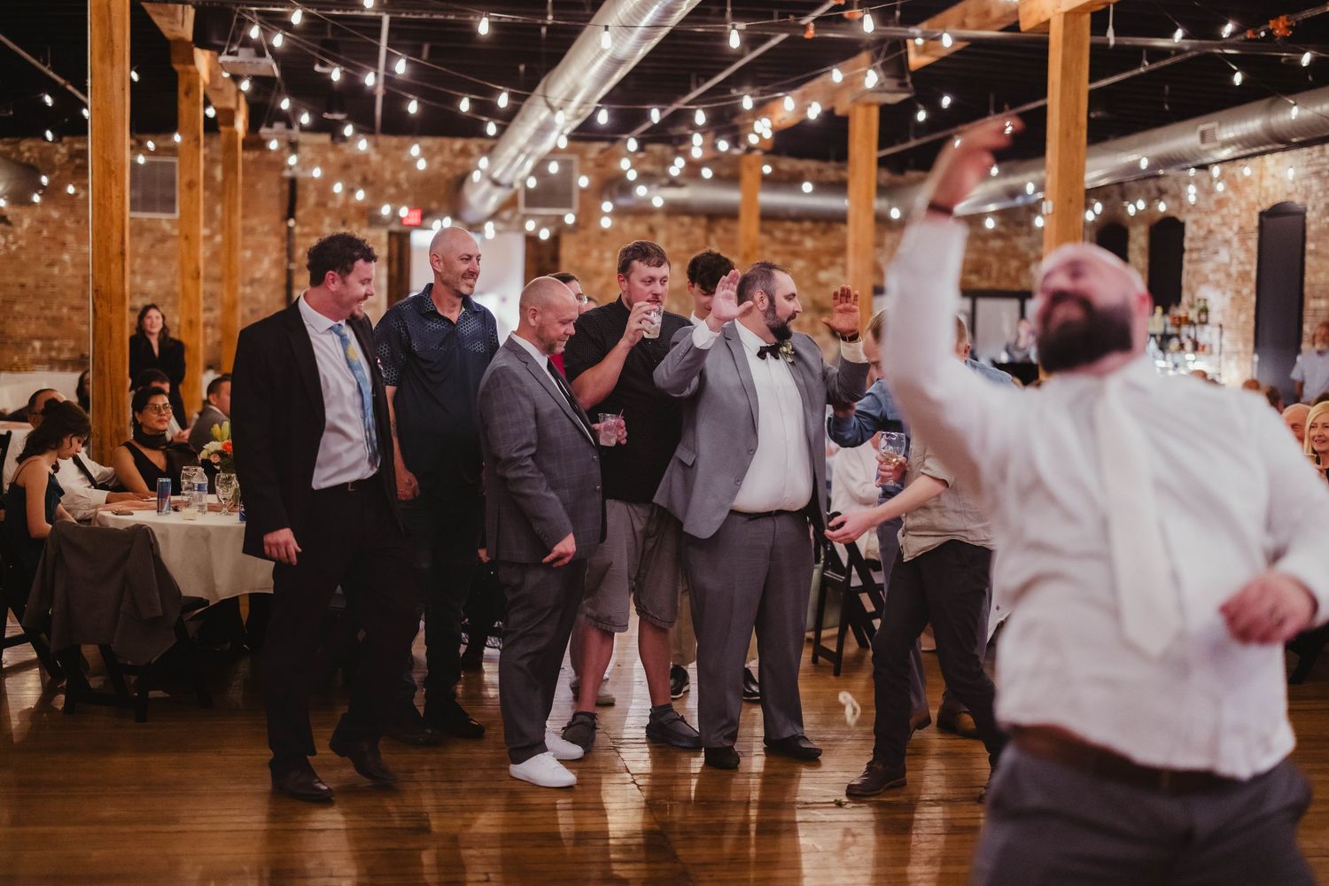 Wedding party dances enthusiastically in a rustic venue illuminated by overhead string lights.
