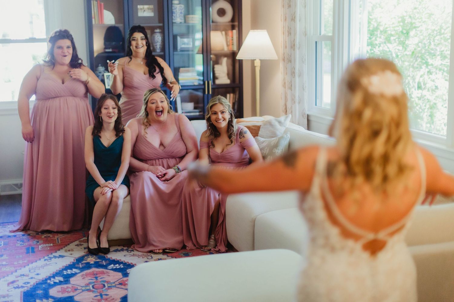 A joyful group moment during wedding preparations in a bright sunlit room with bridesmaids in pink dresses.