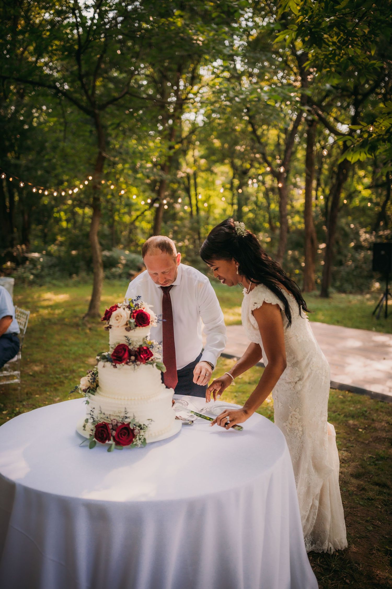 Newlyweds signing marriage documents at a round table outdoors with string lights and trees in the background.
