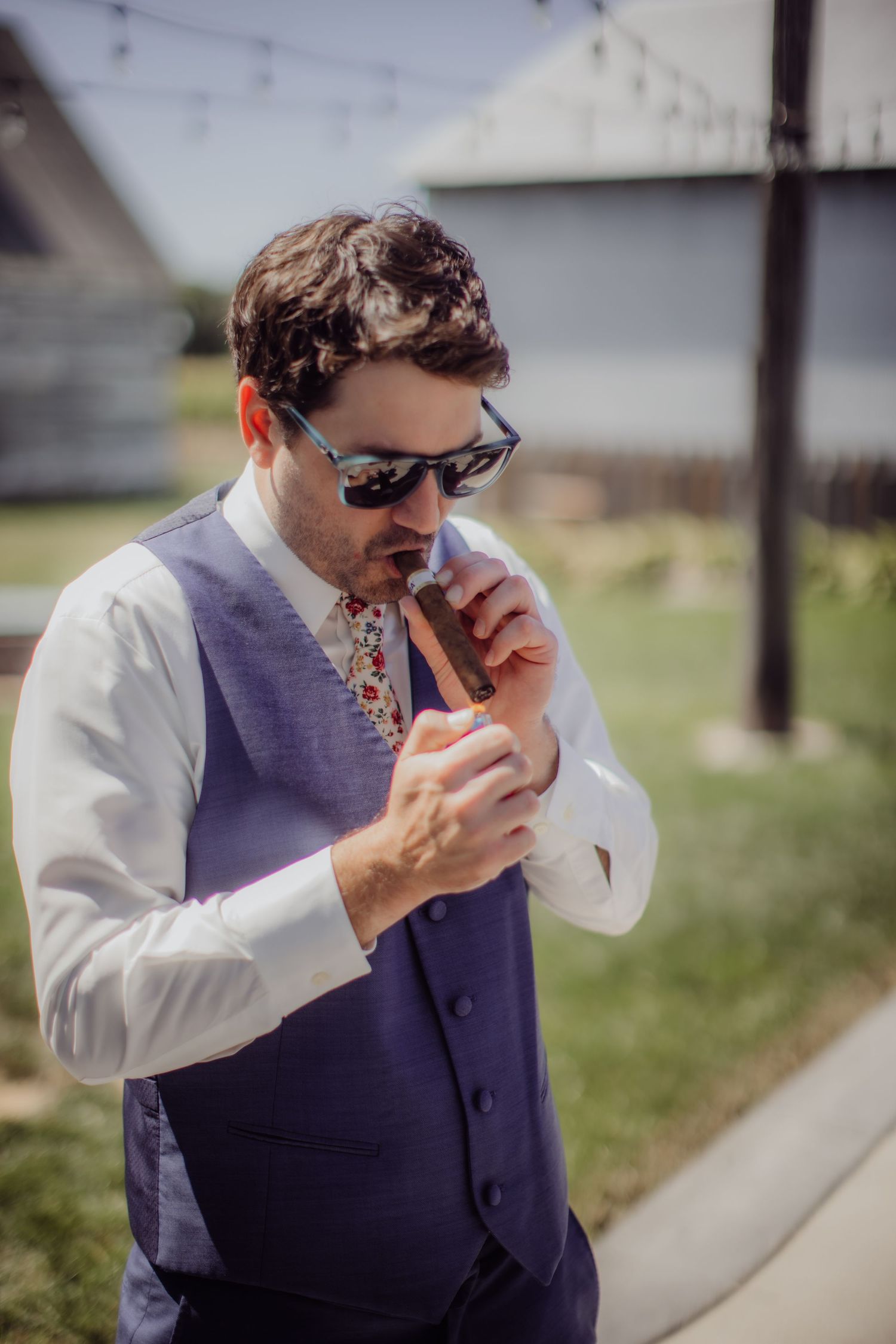 Groomsman in navy vest and sunglasses lights a cigar outside.