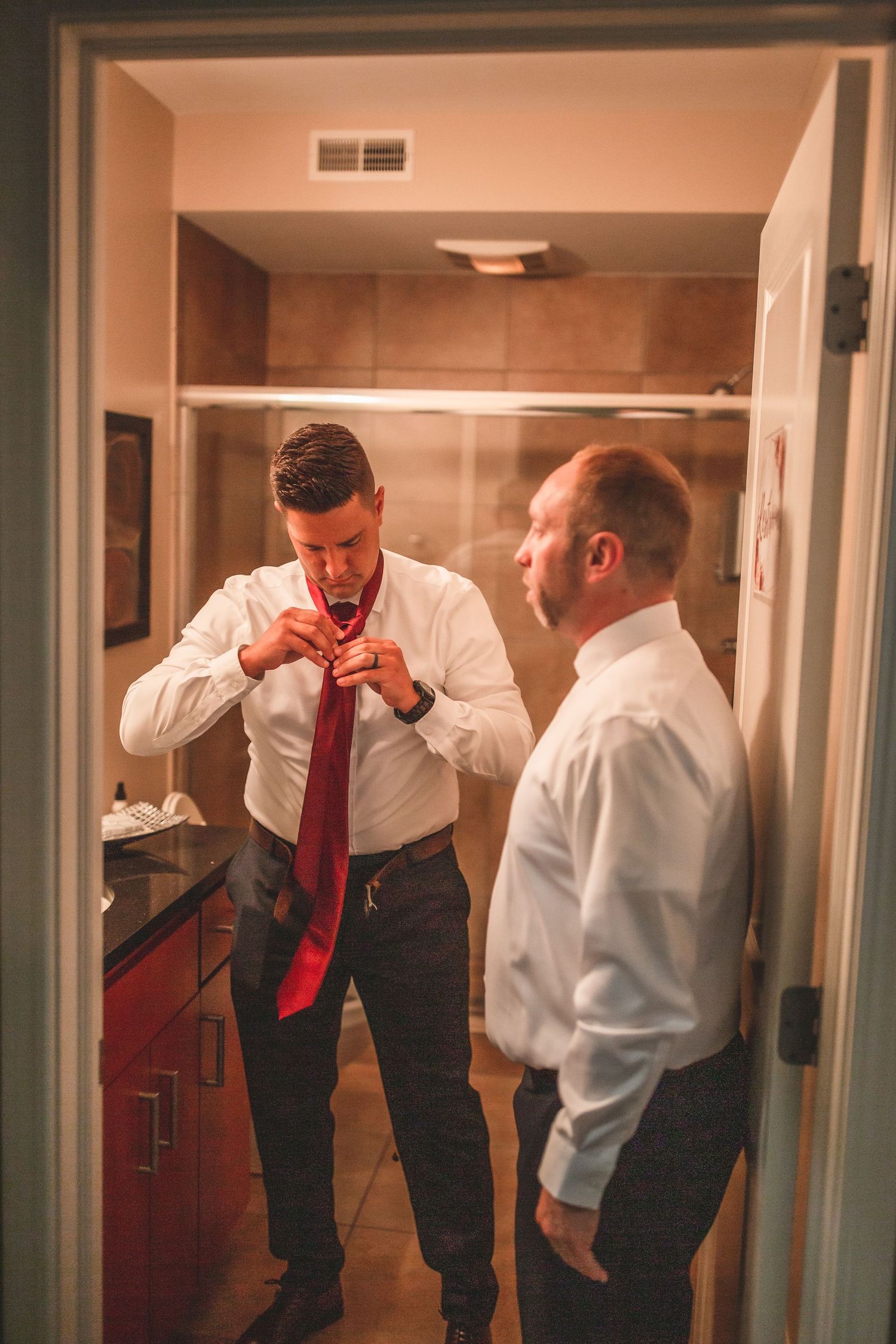 Two men in white dress shirts preparing formal attire in a bathroom, with one adjusting a red tie in the mirror.