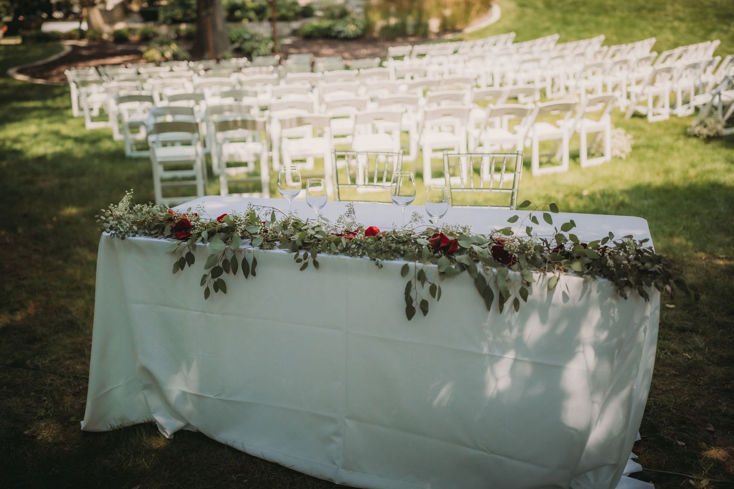 Rows of white ceremony chairs arranged outdoors with floral decorations on a sunny day.