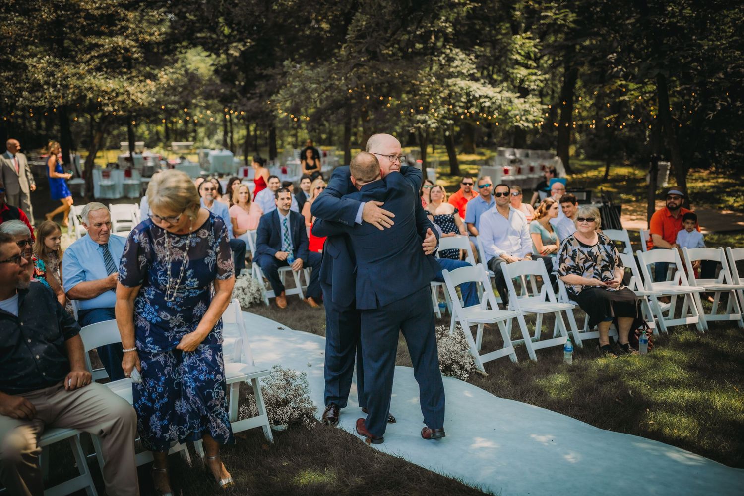 Couples embrace during an outdoor wedding ceremony with white chairs and wooded backdrop.