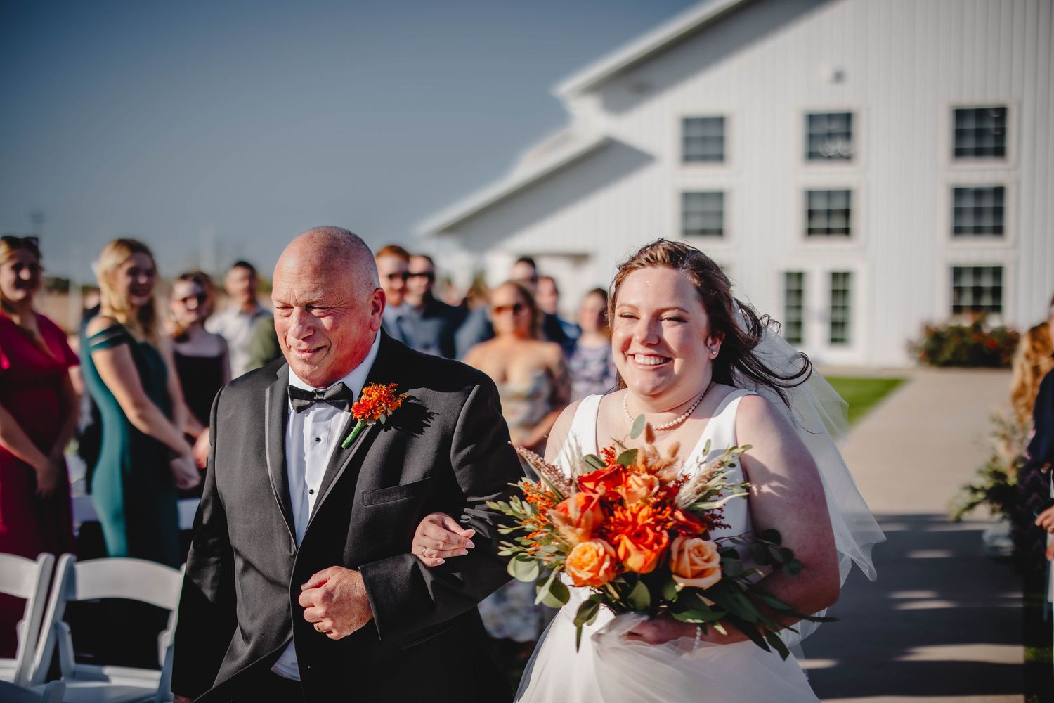 Wedding processional at a white barn venue featuring vibrant orange autumn bouquet.