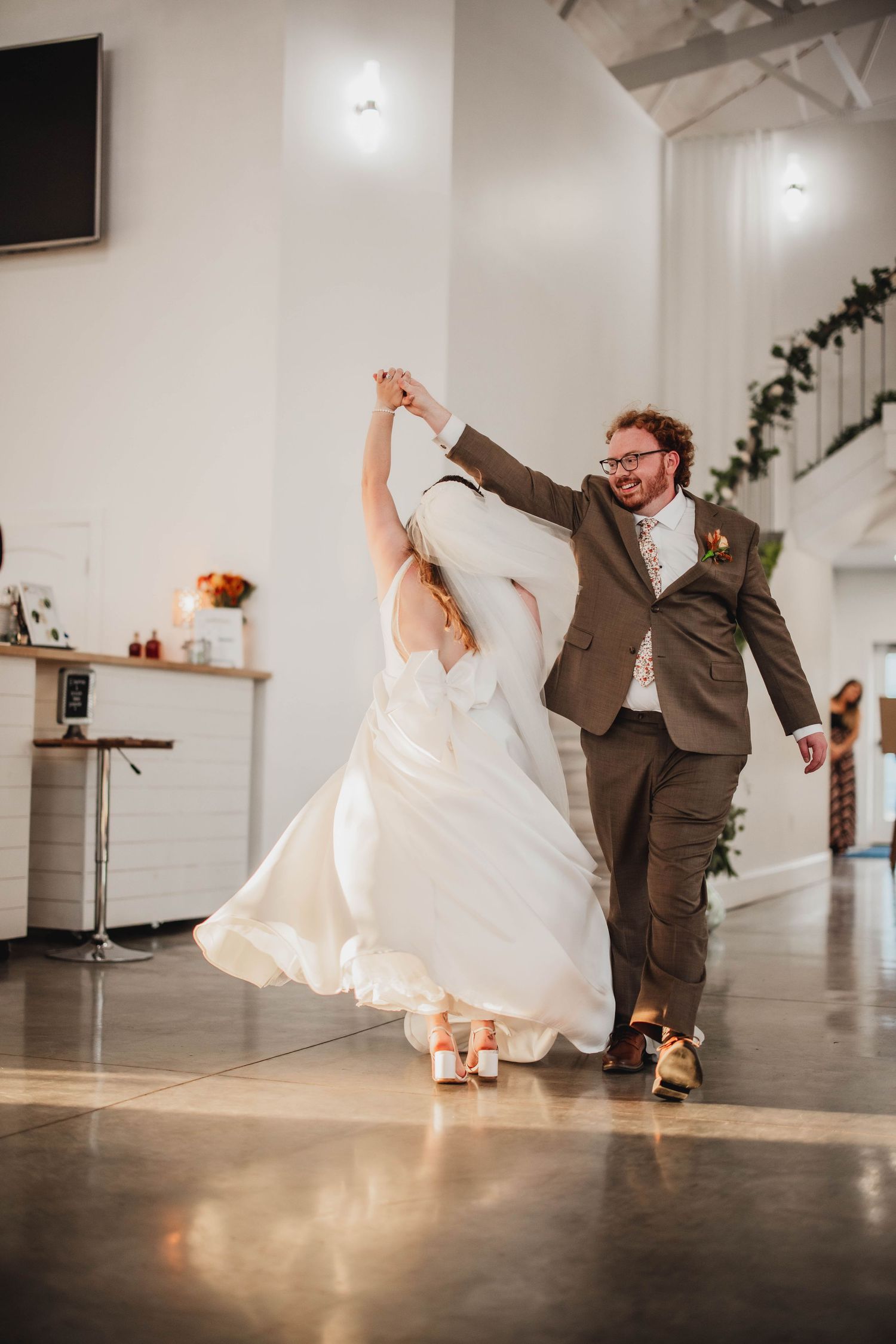 Bride in flowing white gown twirls with groom in brown suit on polished concrete reception floor.