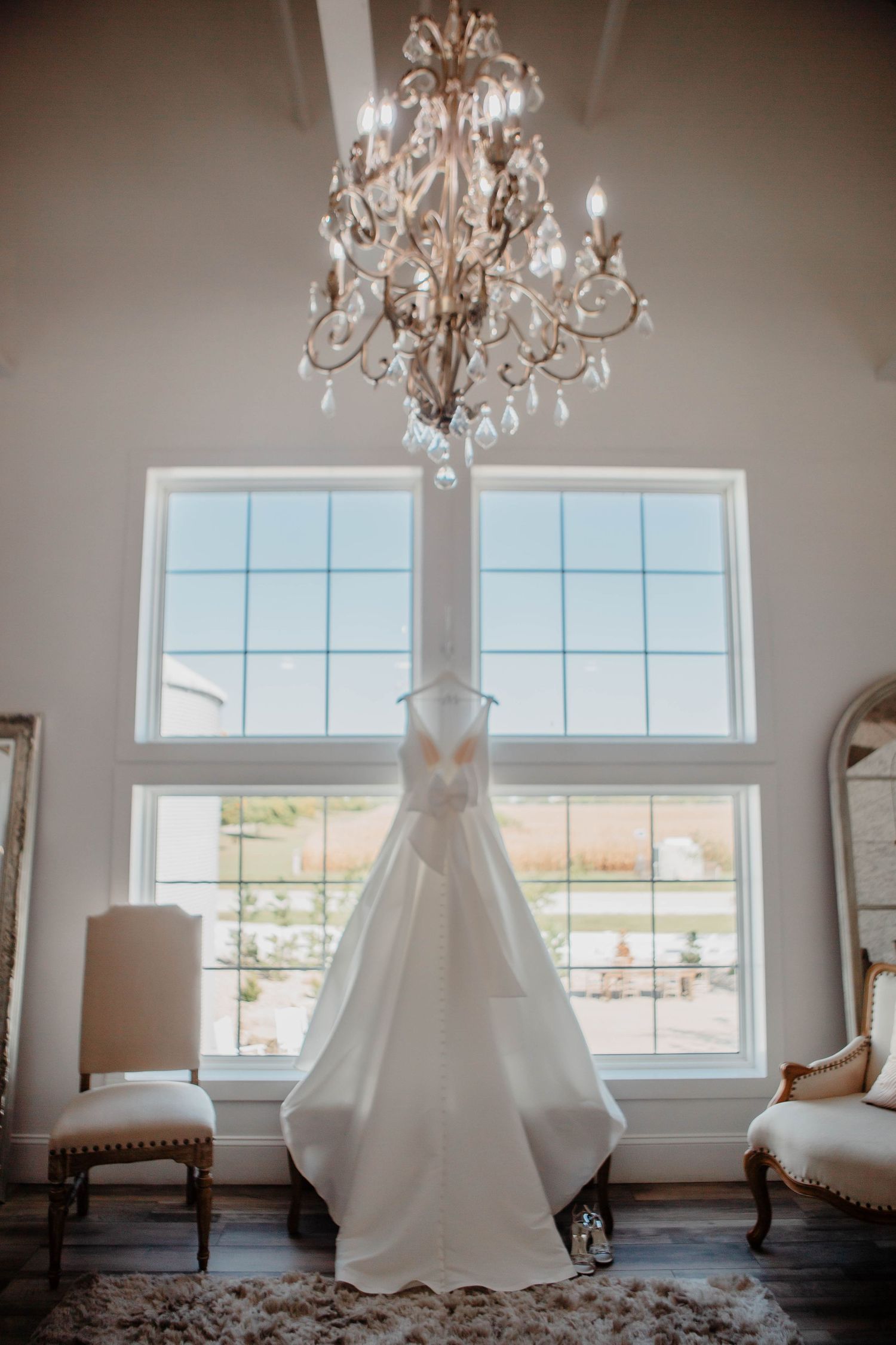 Elegant white wedding dress hanging beneath a crystal chandelier in front of large windows.