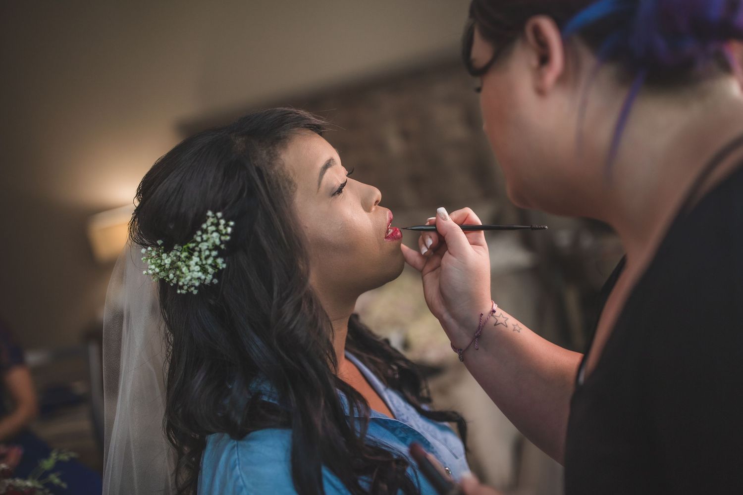 Makeup artist applying lipstick to bride with flowers in her hair.