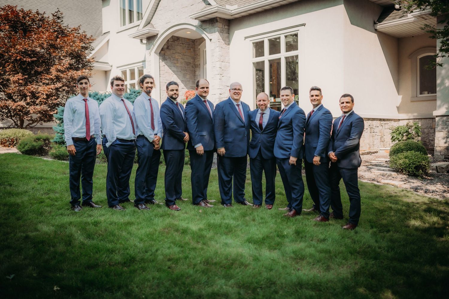 Wedding party group photo featuring navy suits and light blue shirts against cream-colored mansion exterior.