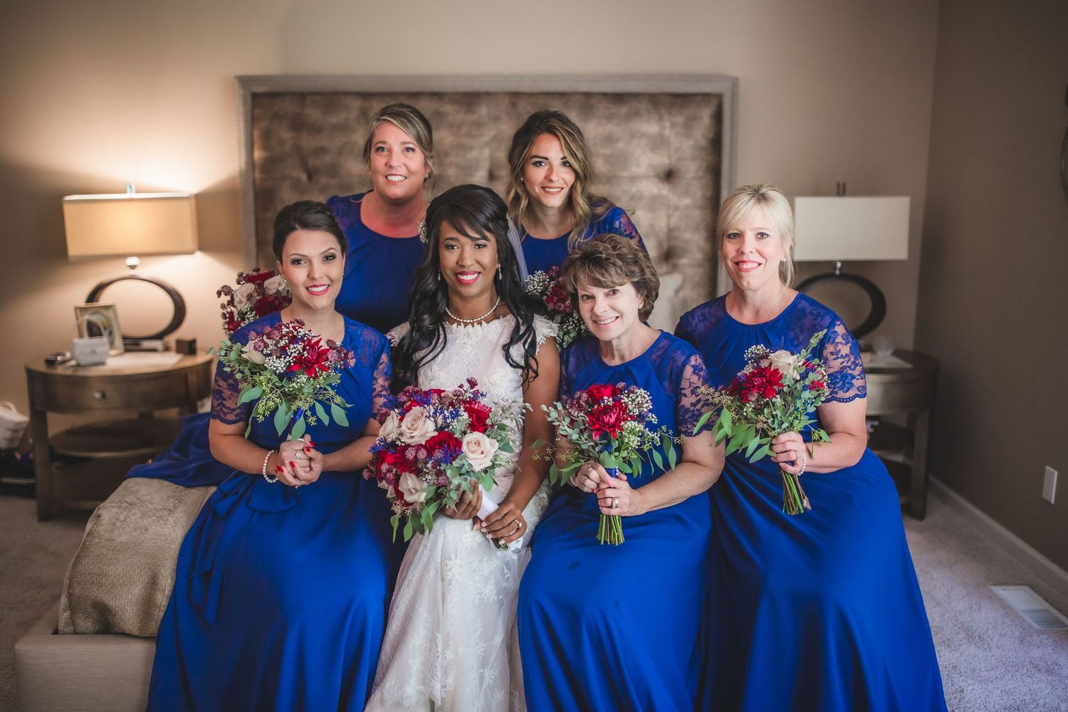 Bridesmaids in royal blue dresses holding vibrant floral bouquets pose on bed.