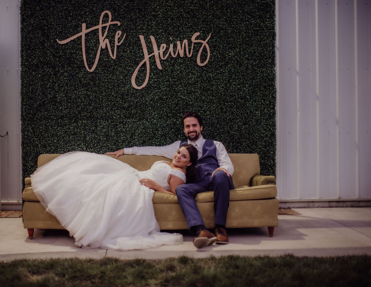 Newlyweds posing casually on a beige couch against an elegant green backdrop.