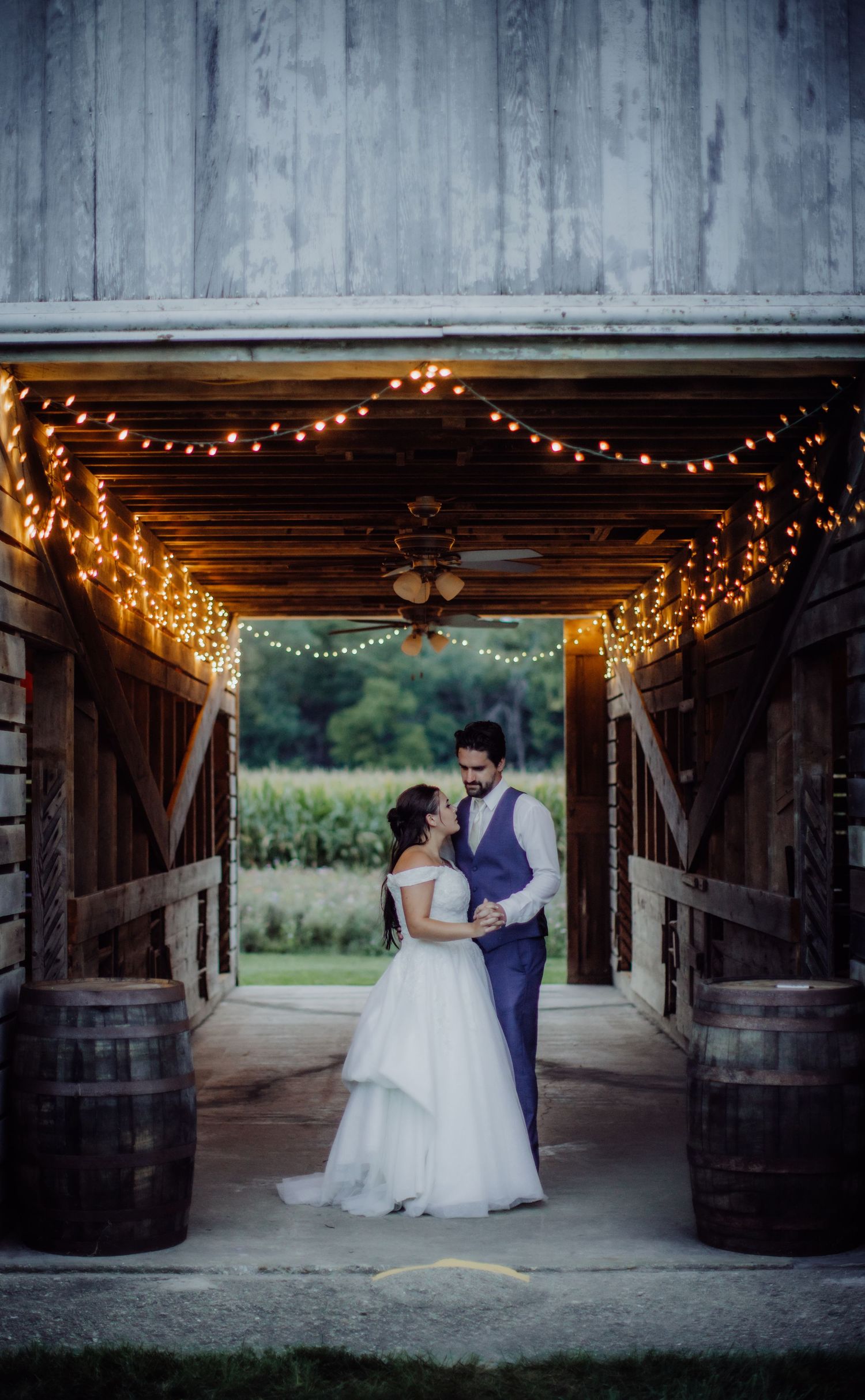 Wedding couple dances in rustic barn venue decorated with warm string lights and wooden barrels.