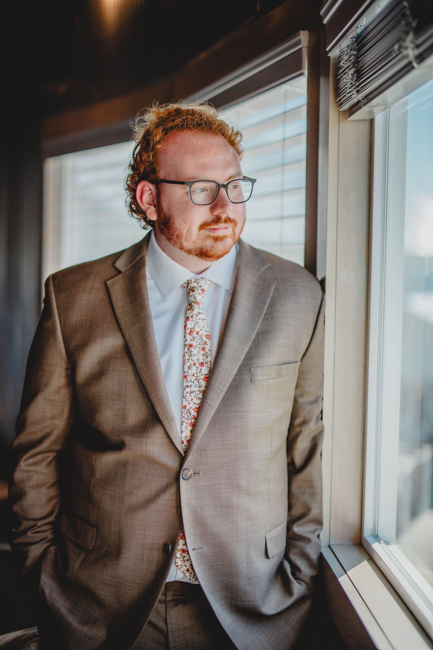Person wearing a grey suit and patterned tie stands by a window in natural lighting.
