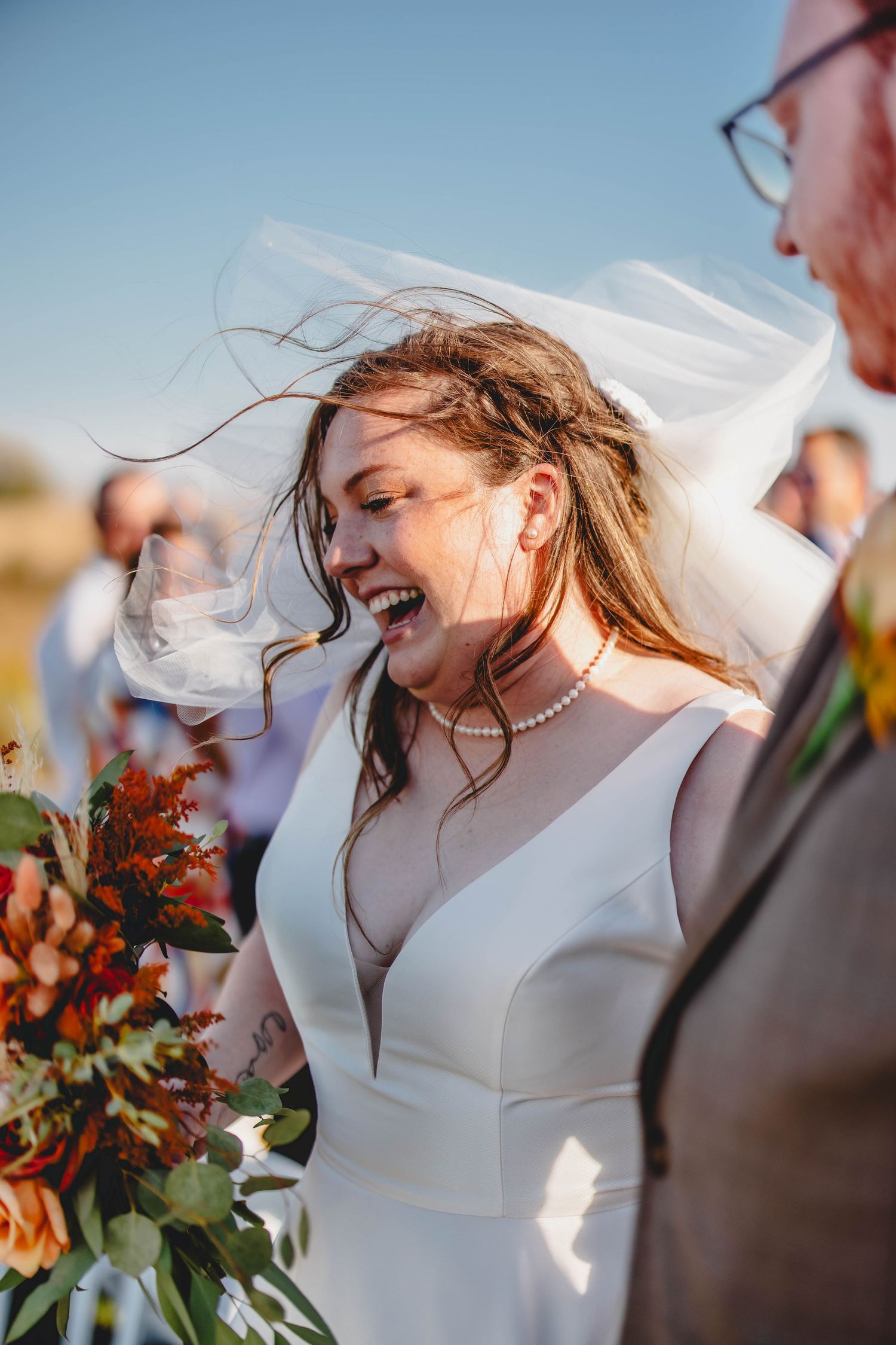 Bride laughs with windswept veil while holding autumn bouquet outdoors.