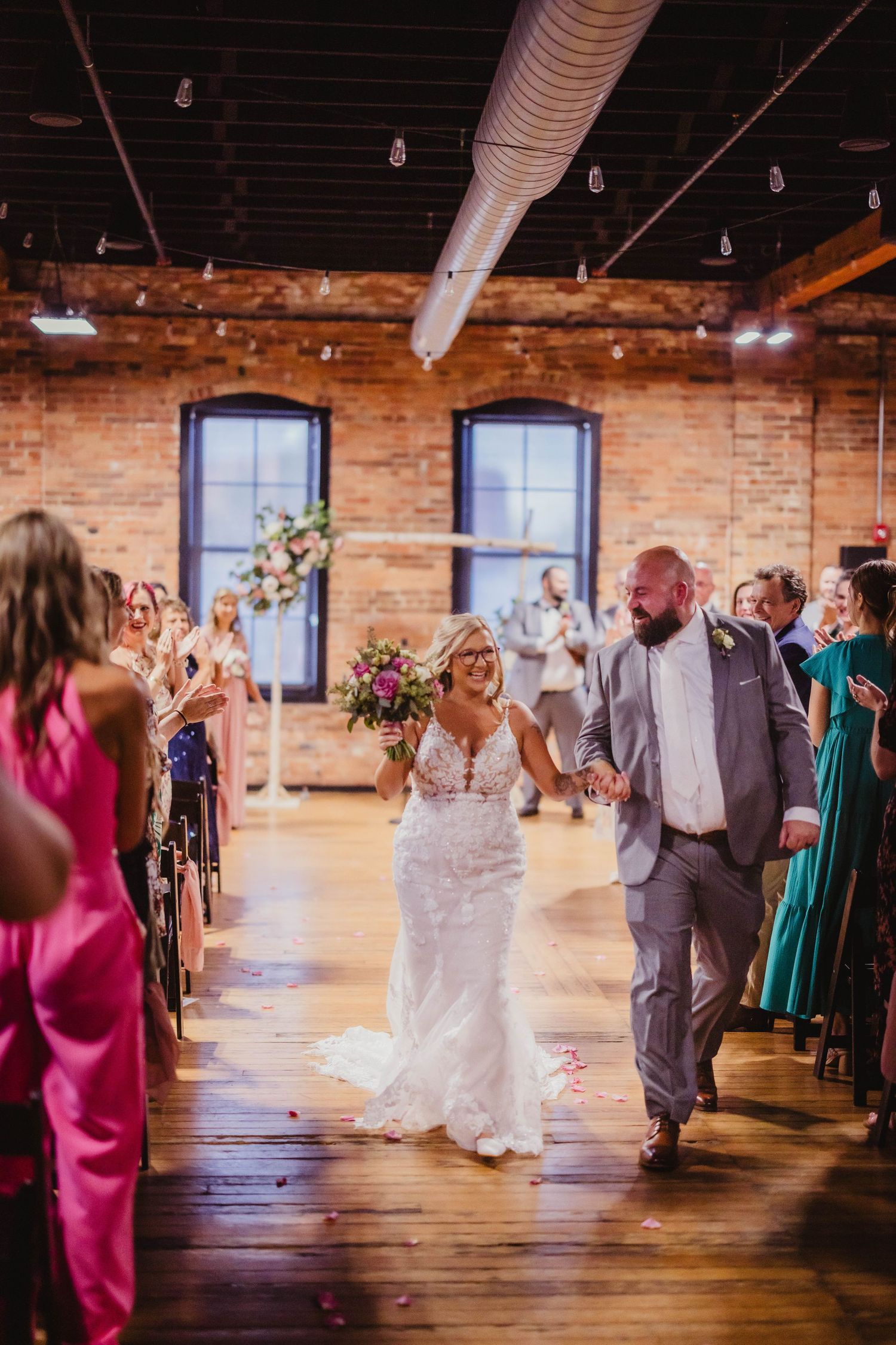 Wedding couple exits ceremony to guest applause in rustic brick venue with hardwood floors and floral arrangements.