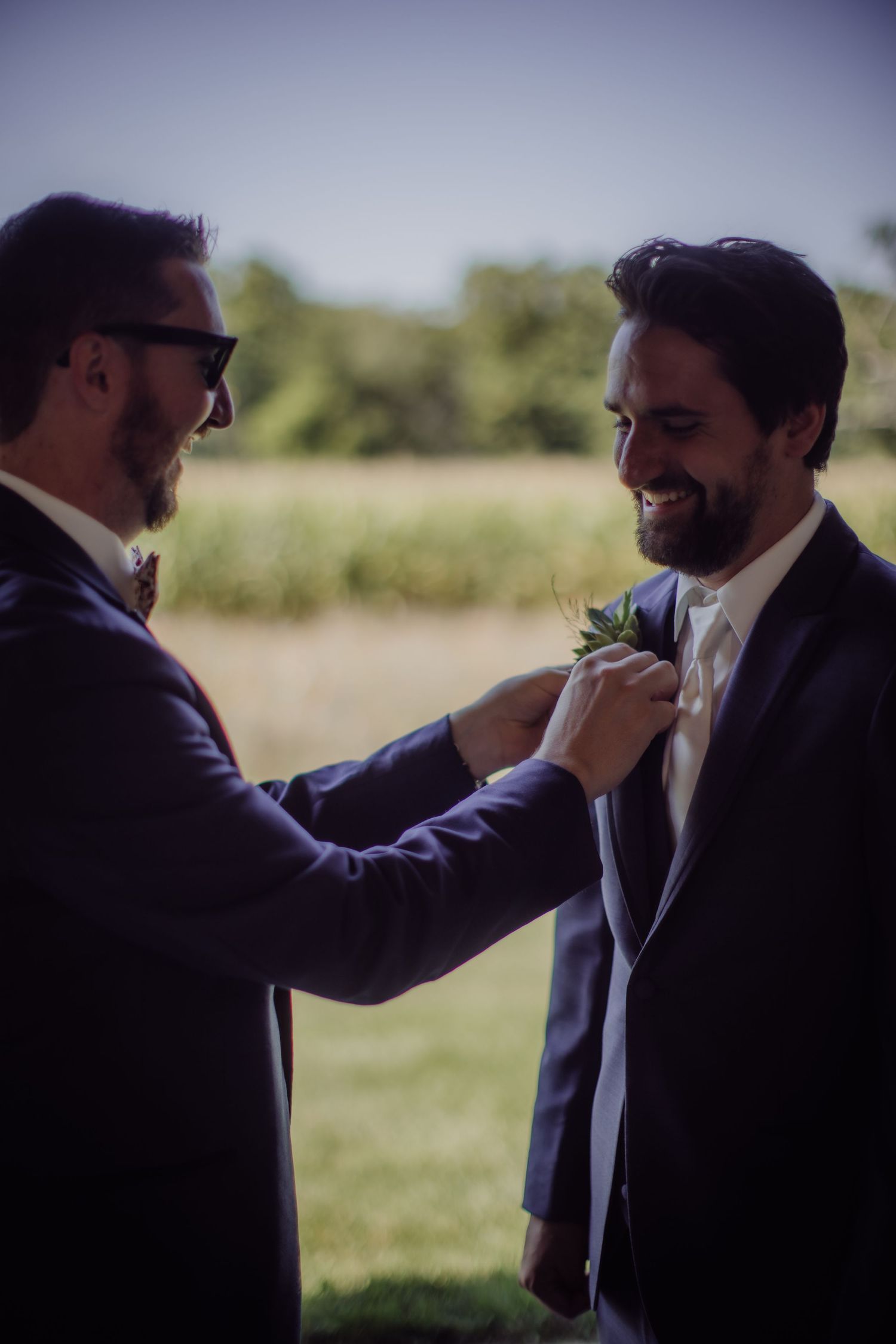 A groomsman adjusts the boutonniere on another's navy suit jacket outdoors.