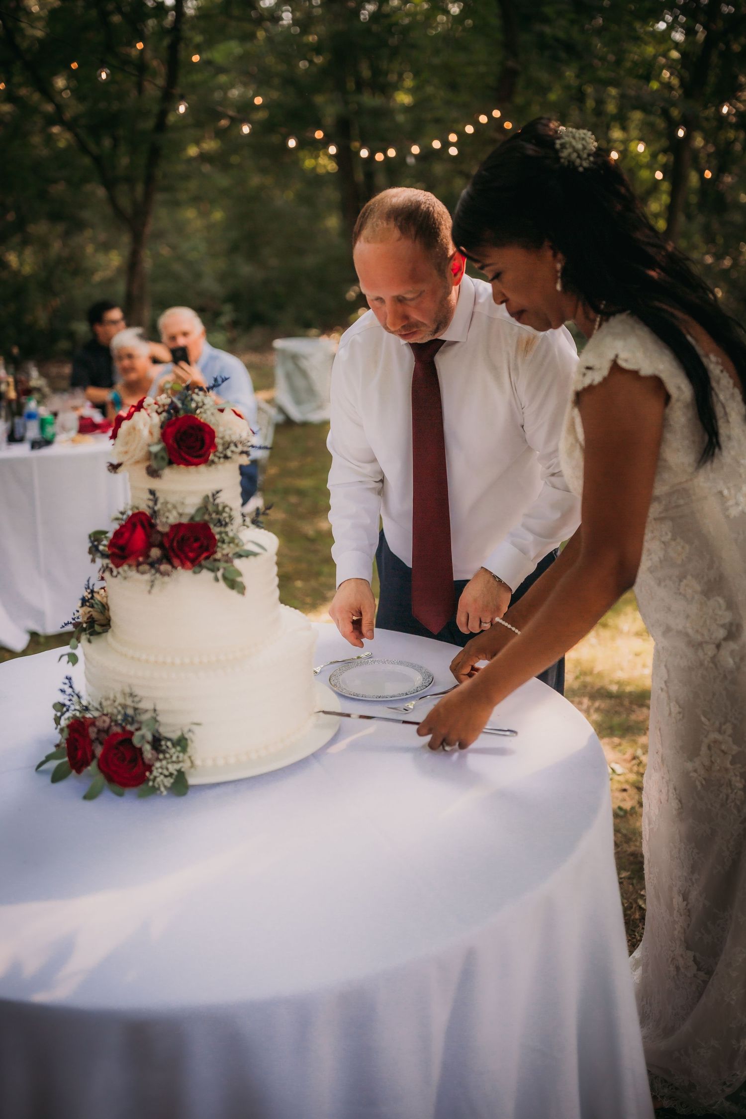 A couple cuts their elegant white wedding cake adorned with red roses during an outdoor garden ceremony.