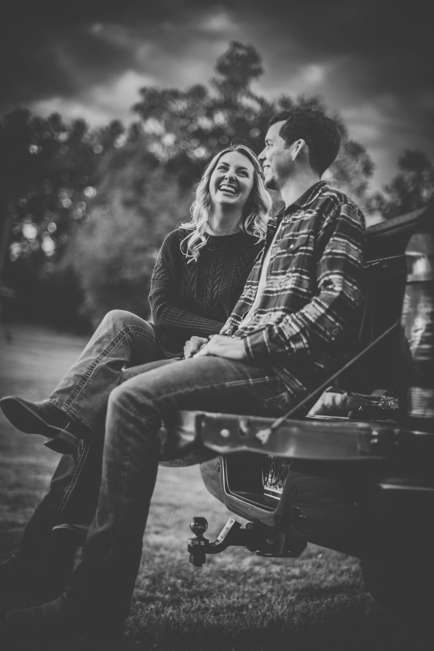 A romantic black and white portrait of a couple sitting and laughing together on the back of a pickup truck.