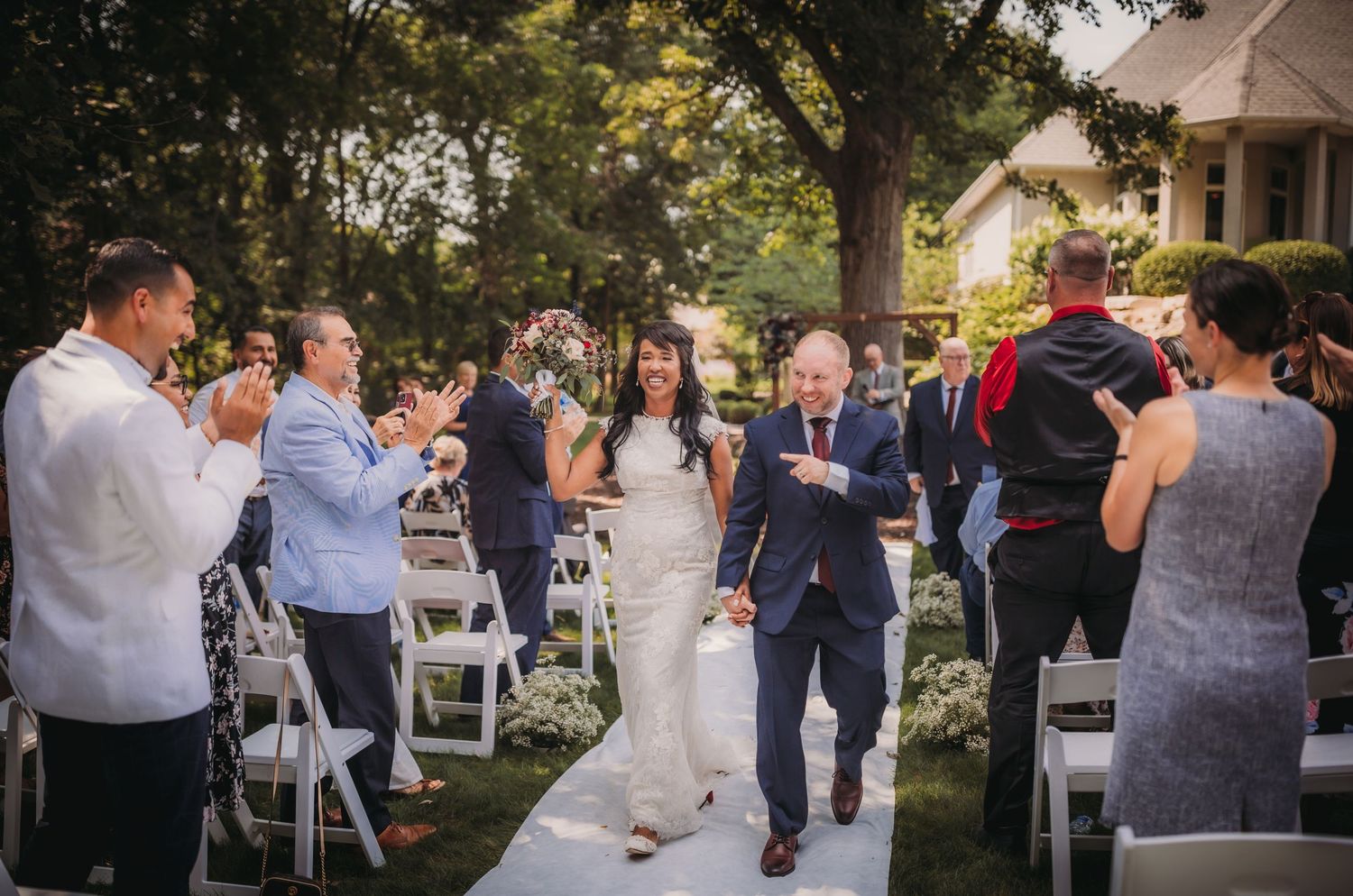 Wedding guests socialize during an outdoor reception beneath towering trees.