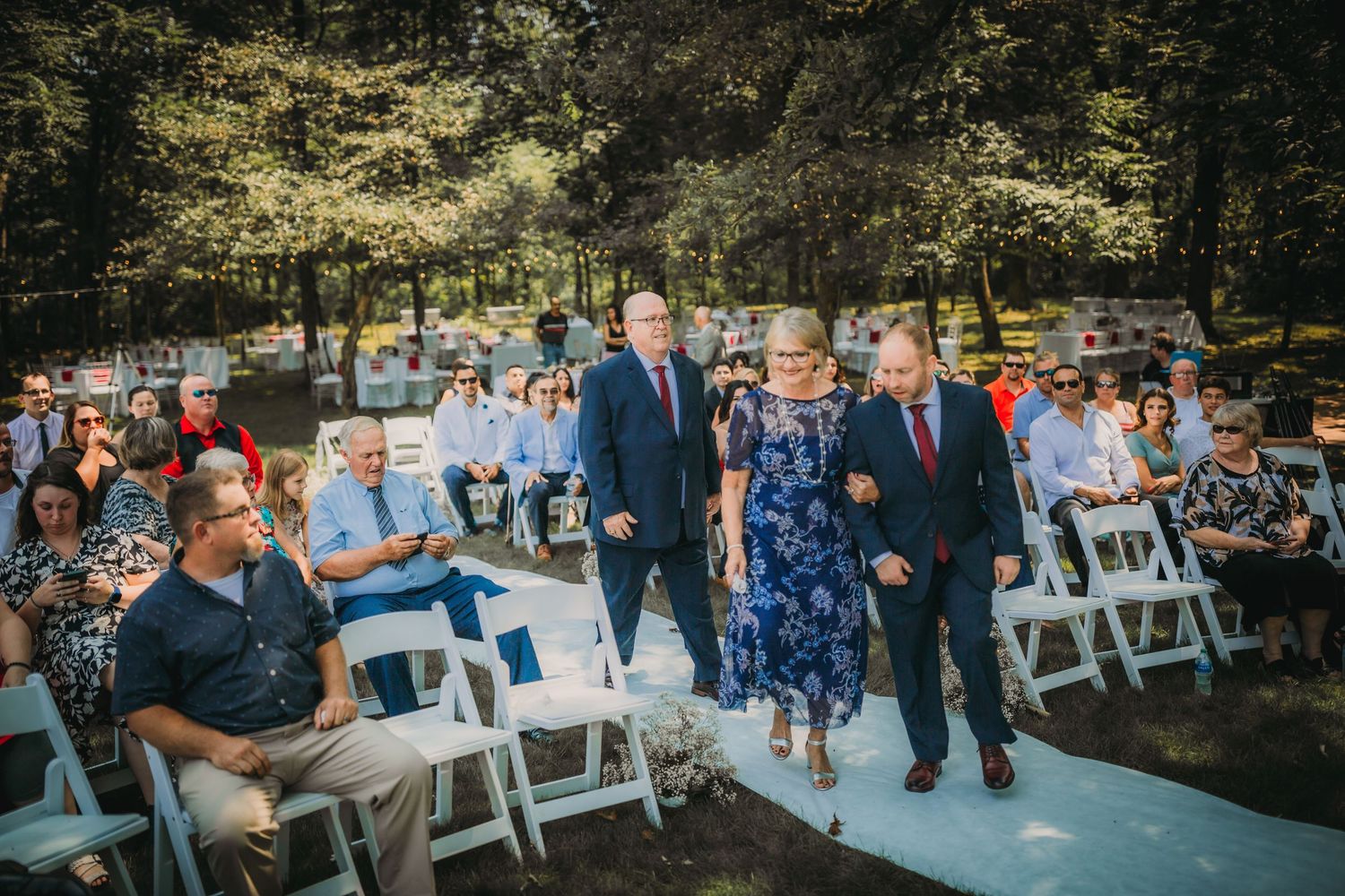 Outdoor wedding ceremony with white chairs and guests gathered in a wooded setting.