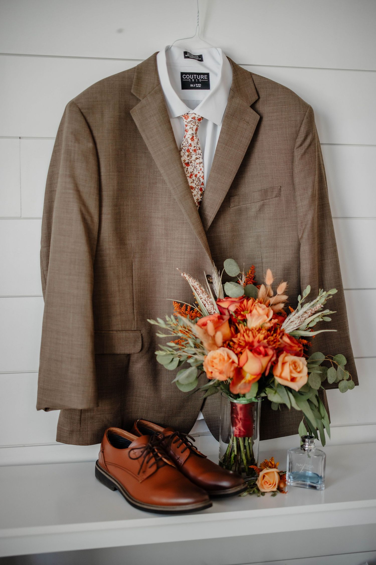 Brown suit jacket with floral tie and orange bouquet displayed next to brown leather shoes.