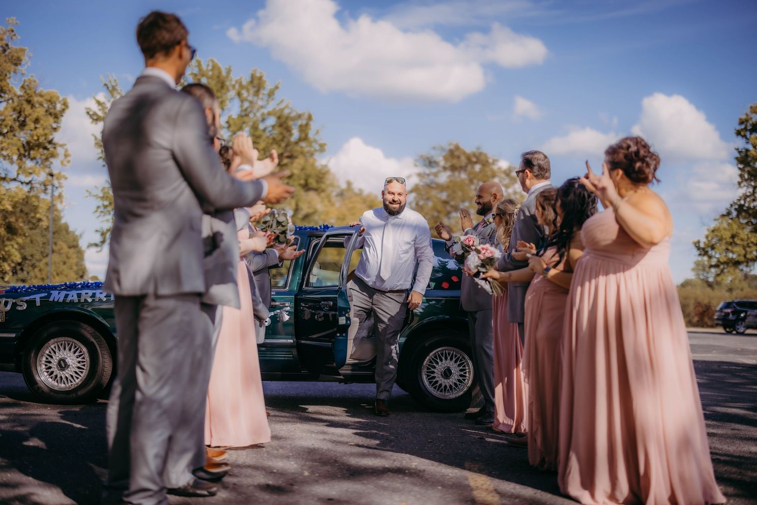 Wedding group stands near classic car outdoors with bridesmaids in pink and groomsmen in gray.