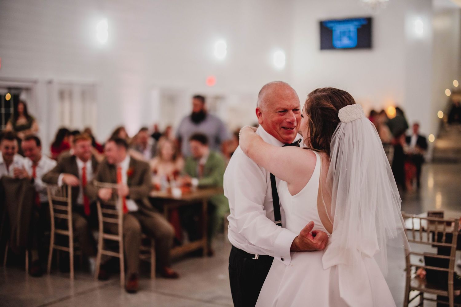 A romantic wedding dance moment captured with reception guests visible in the background.