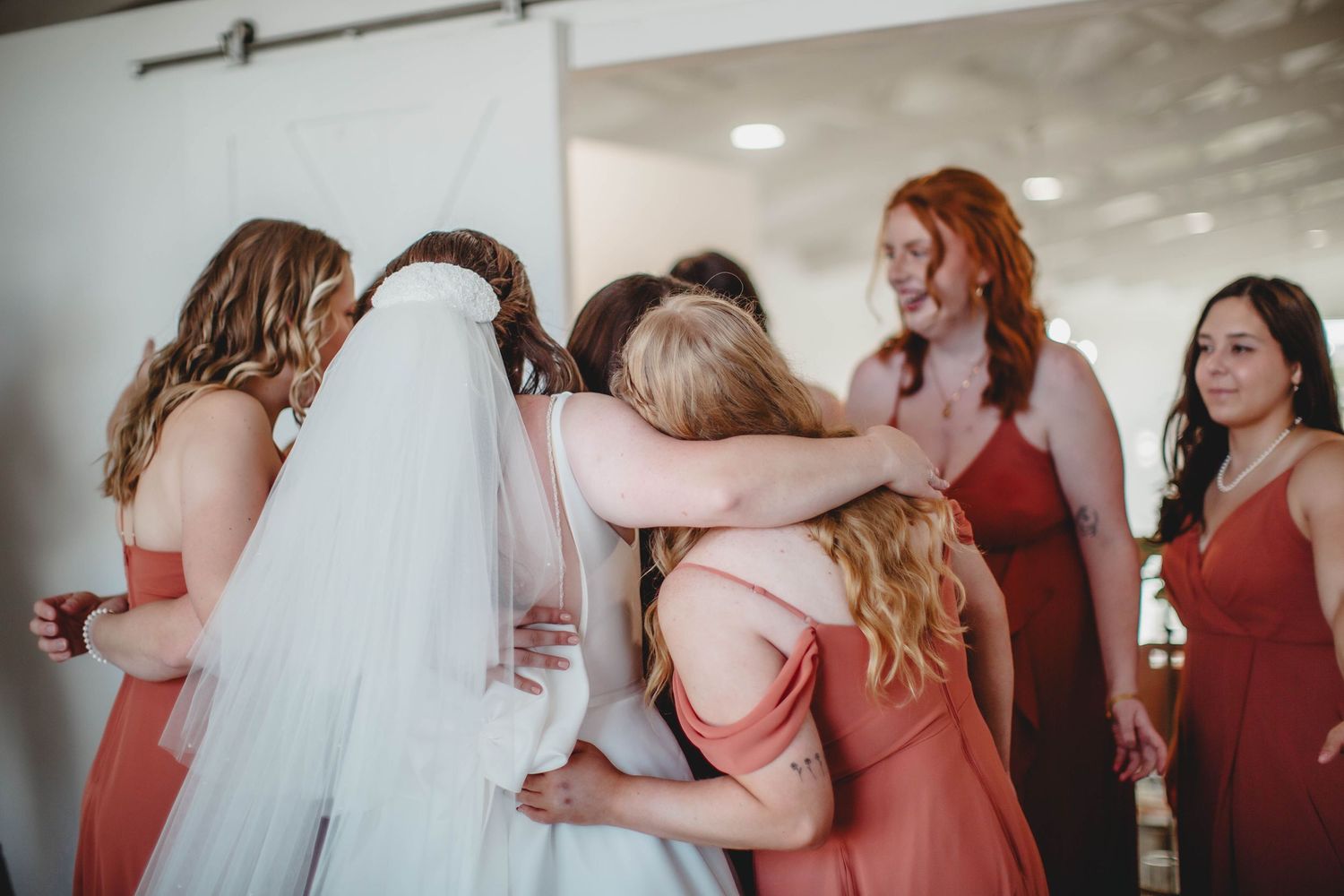 Emotional group hug between bride and bridesmaids wearing coral dresses during wedding preparations.