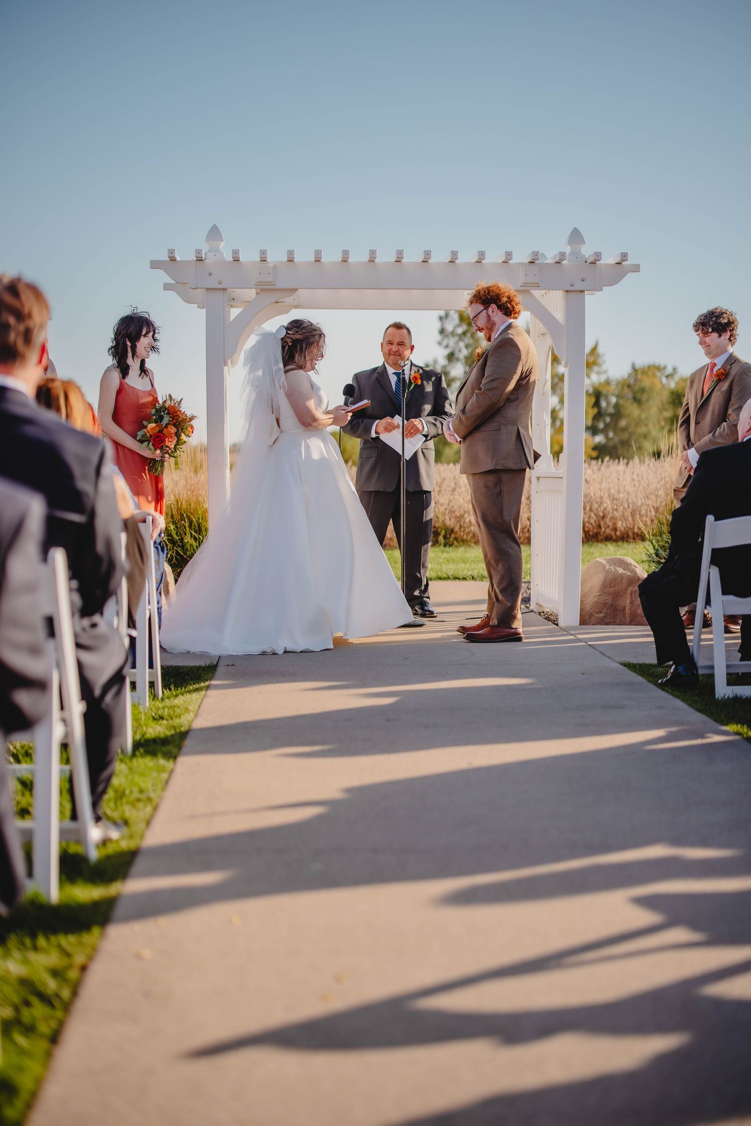 Sunlit outdoor wedding ceremony with elegant white pergola and lined white chairs.