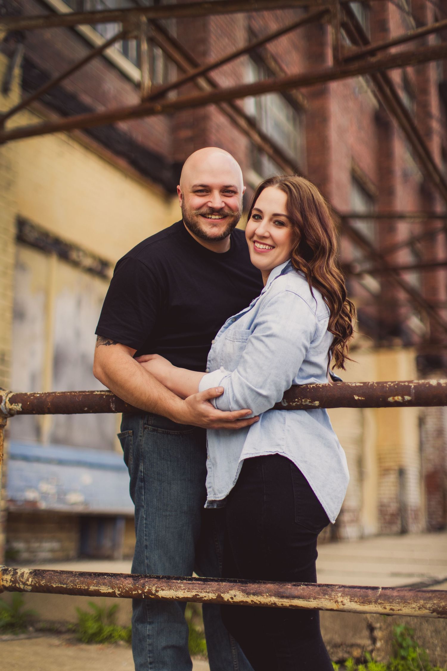 A couple embraces while posing in an industrial setting with metal beams and brick walls in the background.
