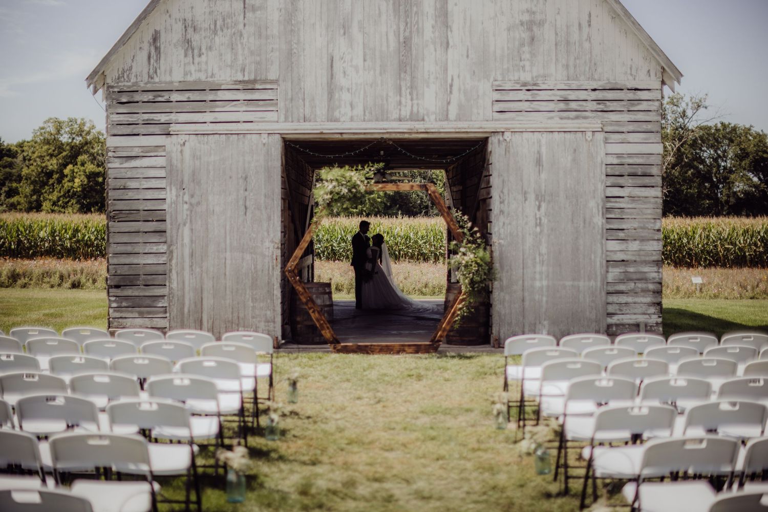 Rustic barn wedding ceremony setup with white chairs leading to wooden barn entrance.
