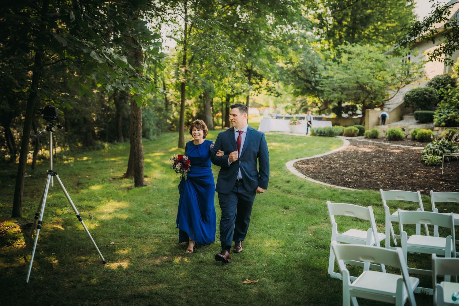 Wedding party members walk together on a winding garden path with white chairs.
