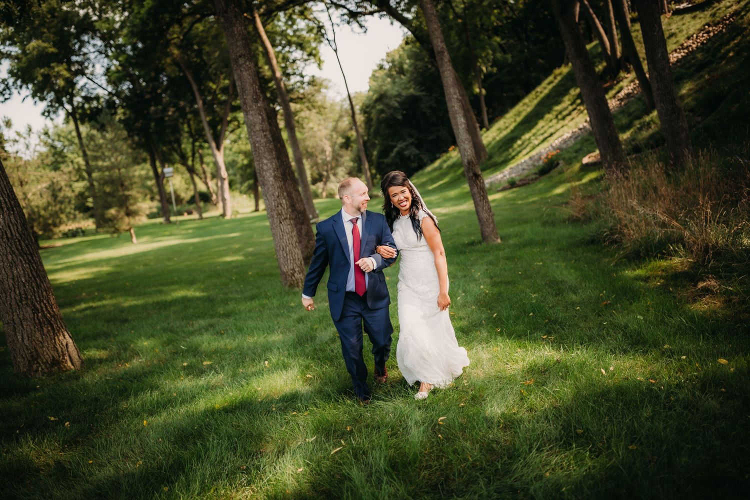 A bride and groom stroll together through a sun-dappled forest setting.