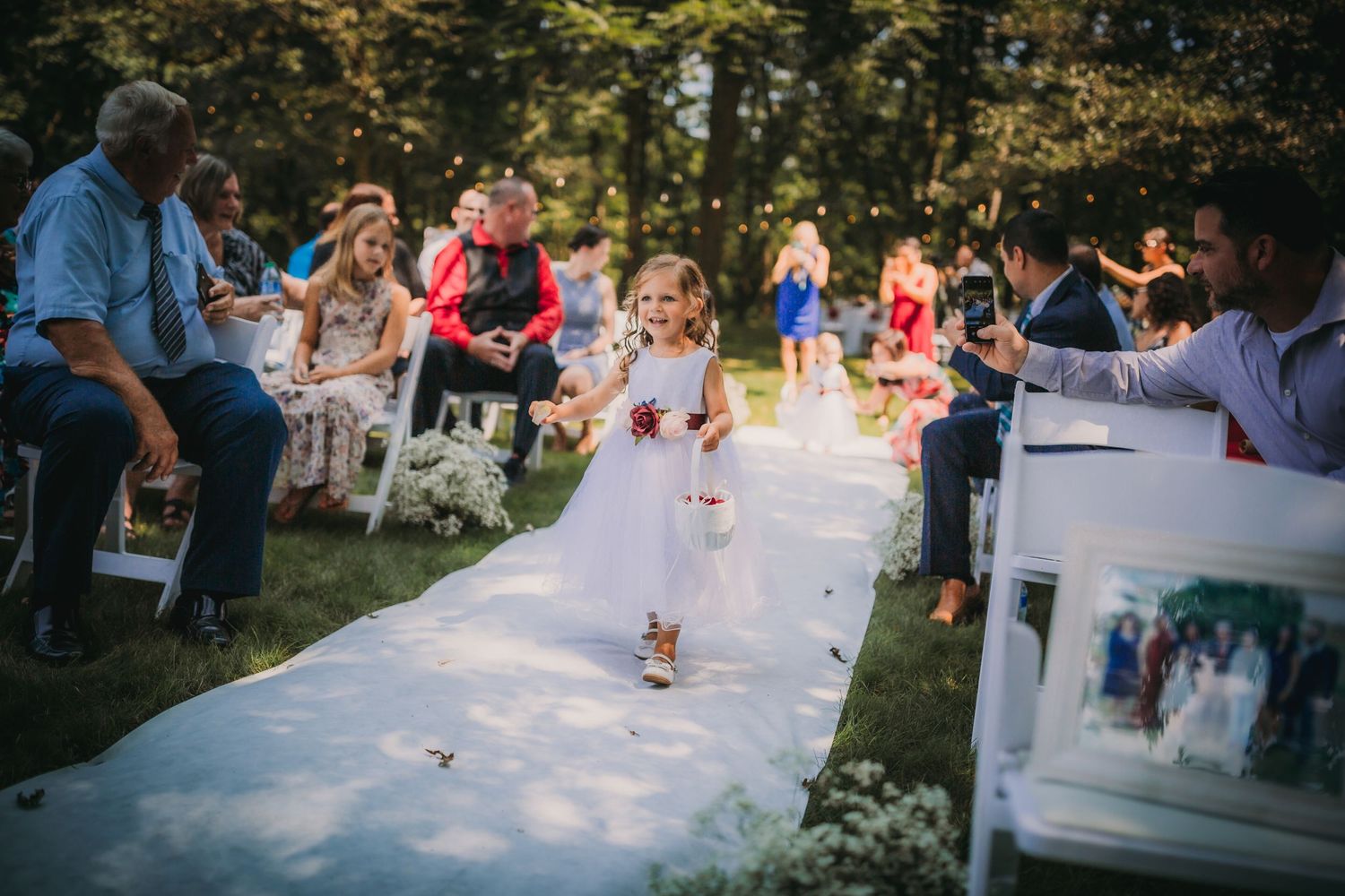 A young attendant carries flowers while walking down a white aisle runner at an outdoor ceremony.