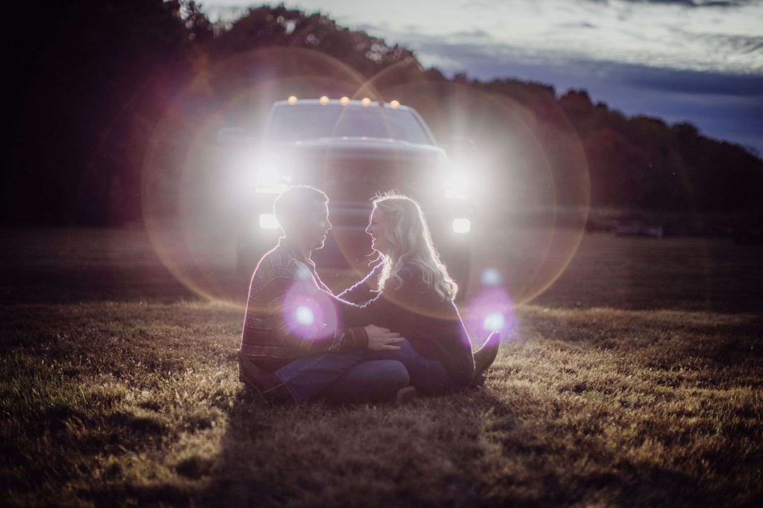 A silhouetted couple sits together in a field with dramatic car lighting creating a romantic atmosphere.
