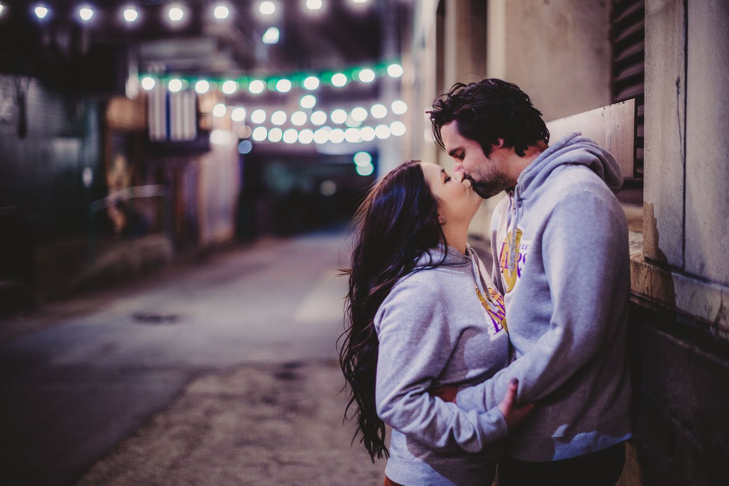 Couple sharing tender moment beneath glowing string lights in city setting.
