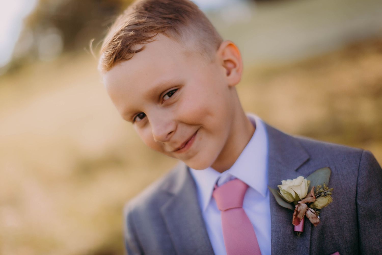 A young attendant in a grey suit and pink tie smiles warmly at a wedding.