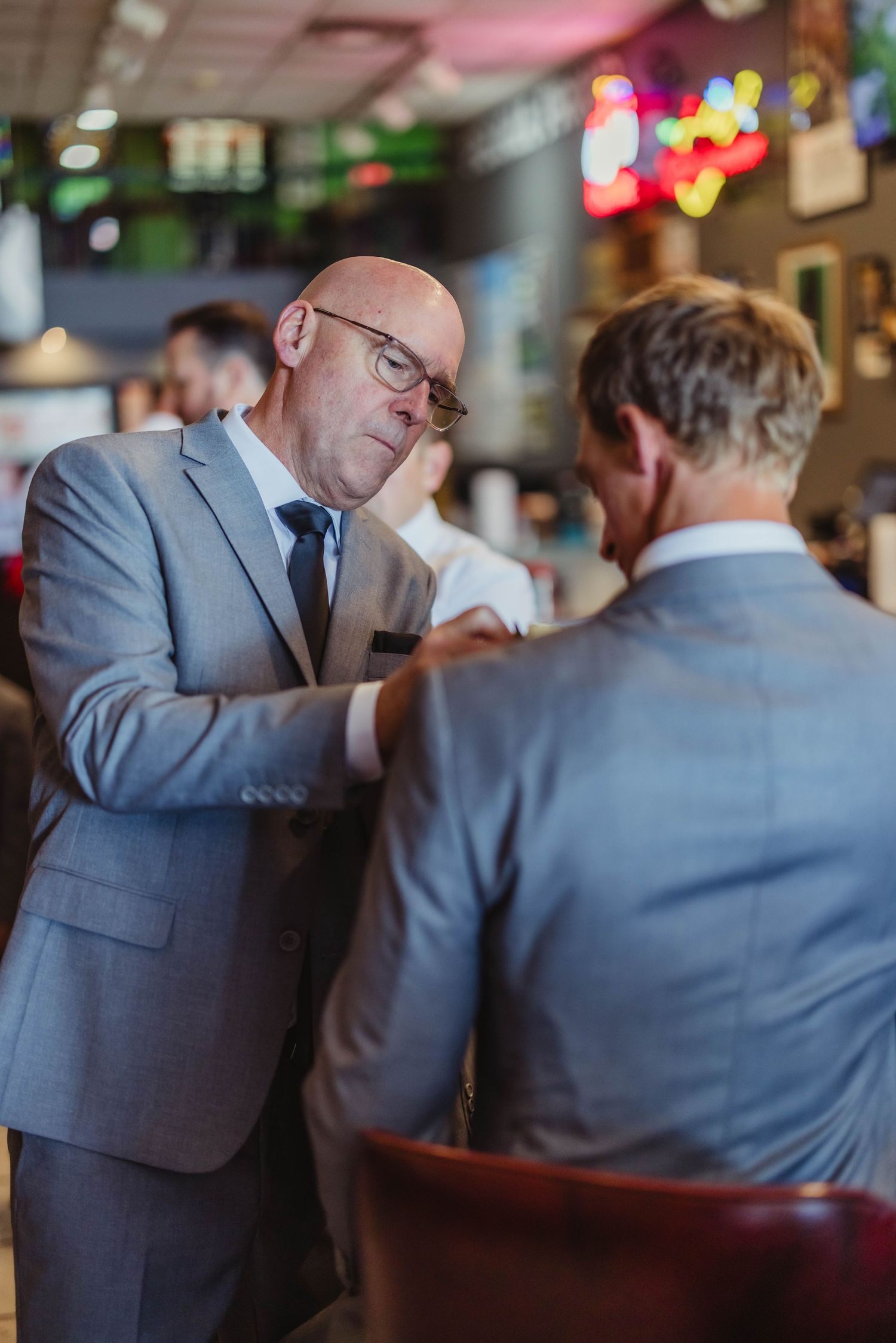 Two people in grey suits help each other with formal attire adjustments in a bar setting.