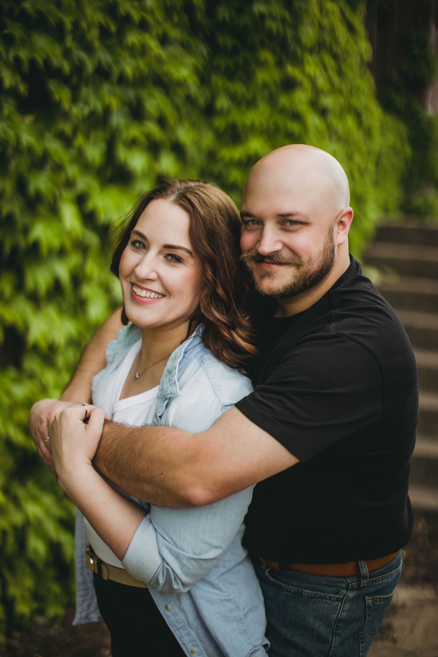 A romantic couple embraces while standing against a lush ivy wall background during an outdoor photoshoot.